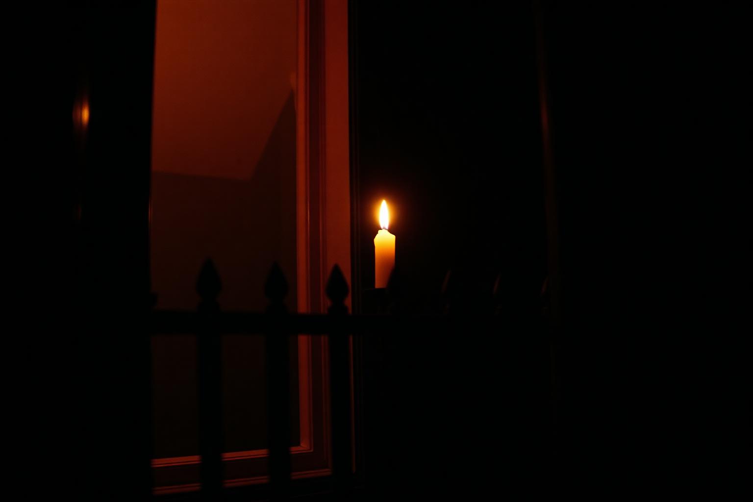 A single lit candle glowing in a New Orleans shotgun-house window at night, seen through wrought-iron porch railings